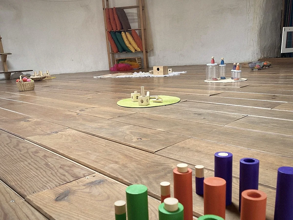 Una sala de Nido preparada: suelo de madera, cestas y bandejas con material de juego, telas de colores y una escalera con cojines