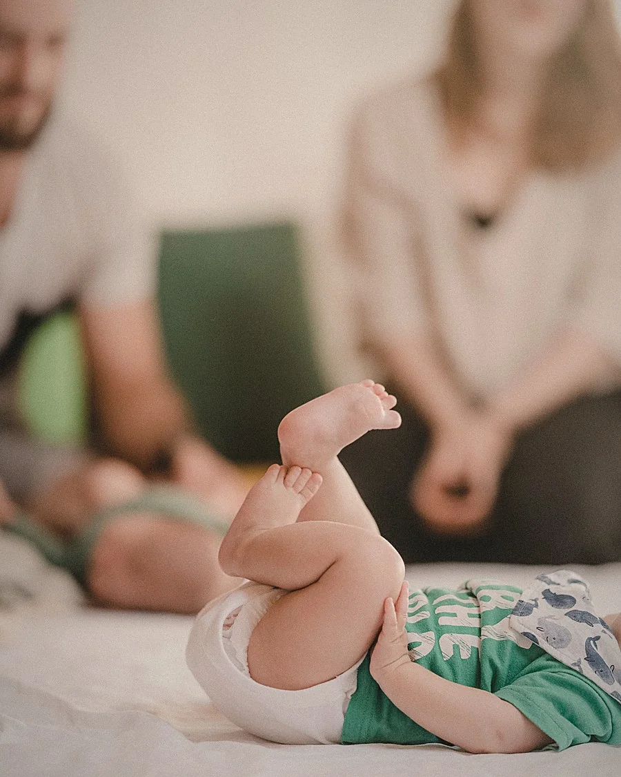 Un bebé tumbado boca arriba durante el cambio de pañal, con su madre y su padre desenfocados detrás