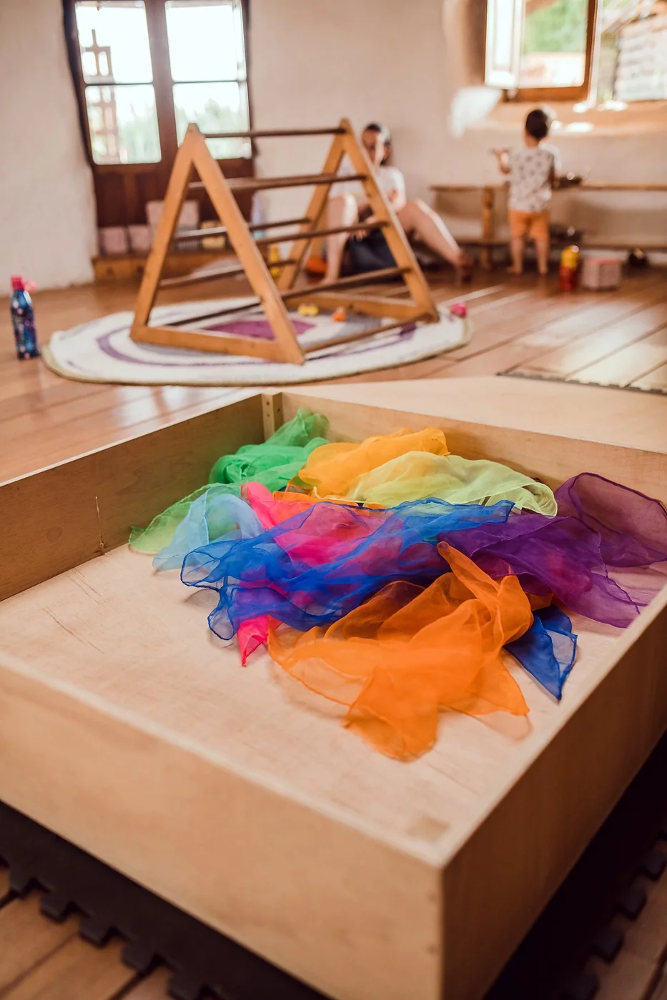 Un triángulo de Pikler en una sala luminosa, con una caja de madera llena de pañuelos de colores